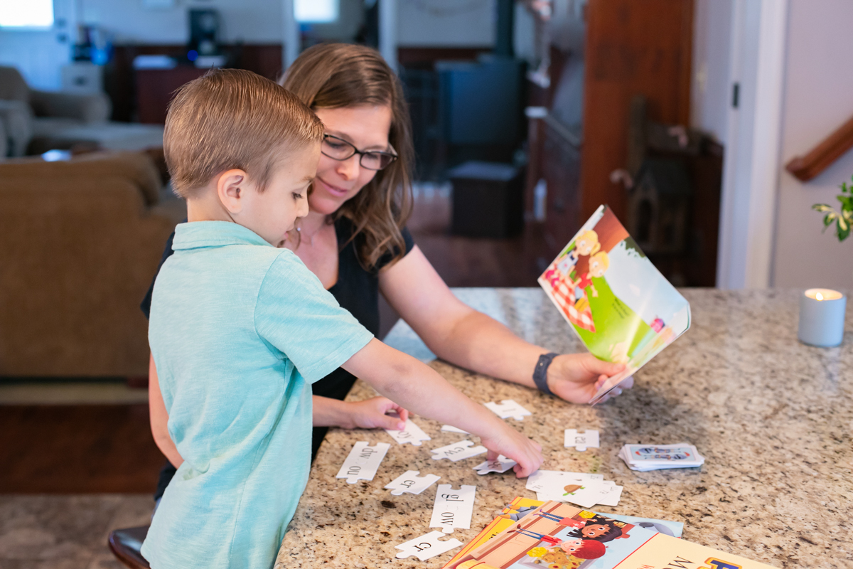 A mother and young son assembling Phonics Museum phonogram puzzle pieces on a kitchen counter while reading a primer together.