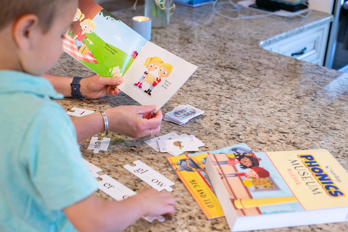 A child fitting Phonics Museum puzzle pieces together beside an open primer and the Phonics Museum workbook.