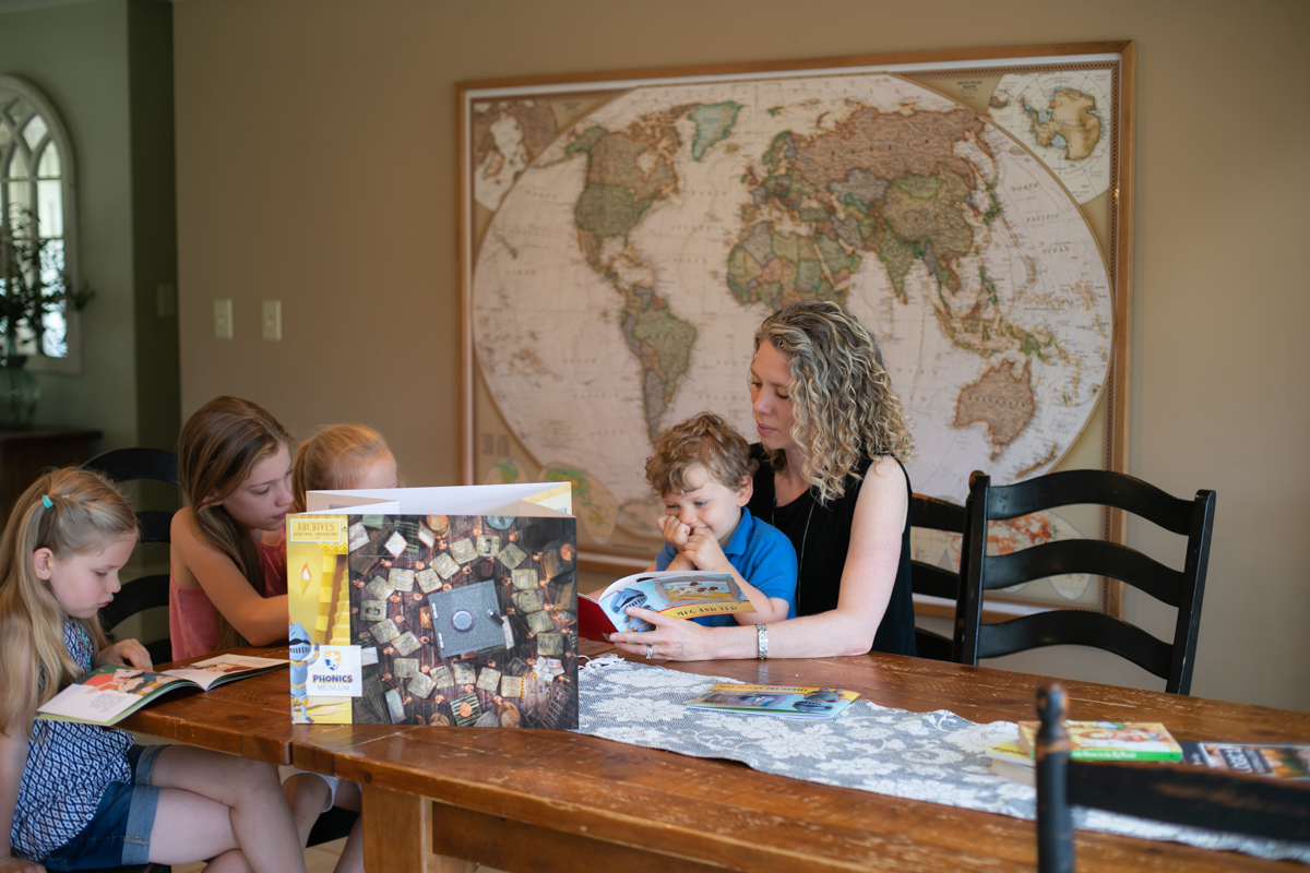 A mother teaching four children of different ages at a dining table with the Phonics Museum pop-up museum board and primers.